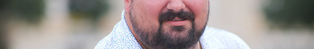 Man with a beard wearing a light blue patterned shirt outdoors
