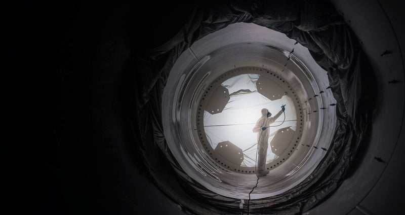 Person in coveralls spraying walls of equipment with sealant or paint.